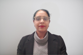 Professional head-and-shoulders portrait of a woman in a dark blazer and patterned blouse, wearing glasses against a light gray background.