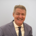 Portrait of a smiling middle-aged man in a light gray suit and navy patterned tie, looking at the camera.