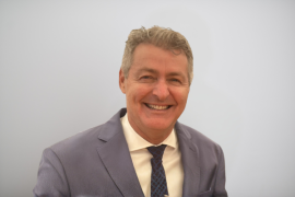 Portrait of a smiling middle-aged man in a light gray suit and navy patterned tie, looking at the camera.
