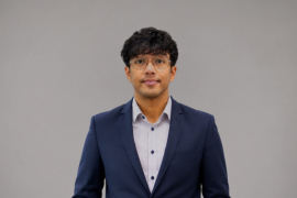 Professional portrait of a man in a navy blazer and light shirt, wearing glasses, against a gray background.
