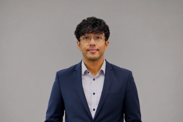 Professional portrait of a man in a navy blazer and light shirt, wearing glasses, against a gray background.